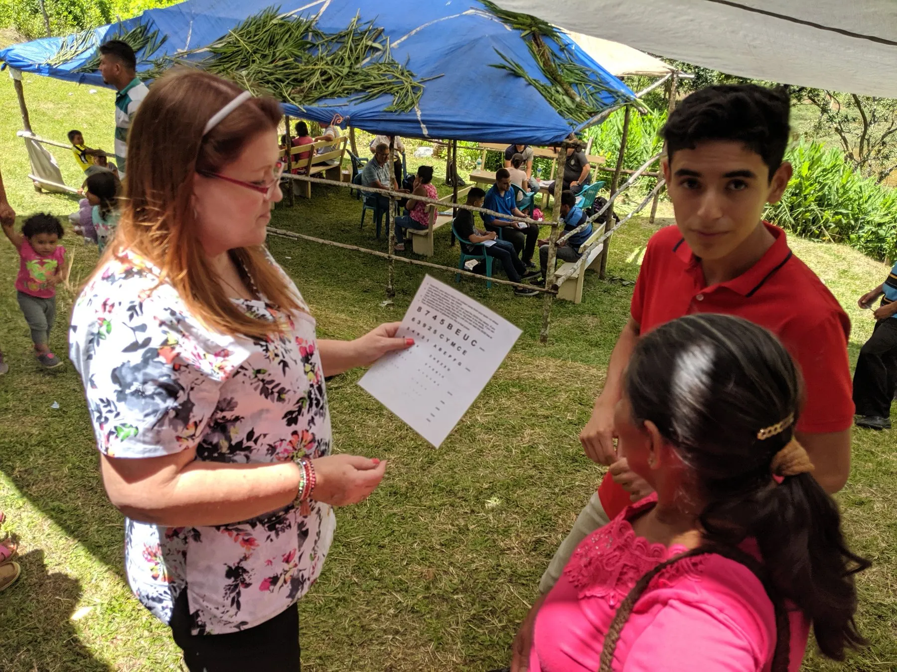 Loring testing a young man's reading with a card at a medical outreach under a blue canopy.